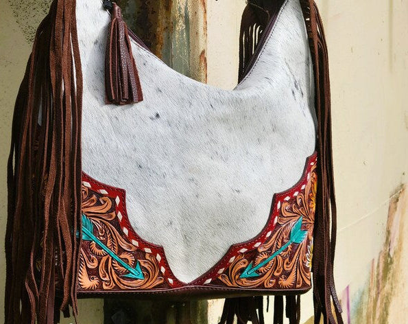 Cowhide leather bag with fringe and intricate designs, featuring 'The Rooster Den Co' branding.